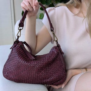 May include: A burgundy-colored woven leather handbag with a shoulder strap. The bag features a zipper closure and brass-colored hardware. The woven design gives the bag a textured appearance. The bag is held by a person.