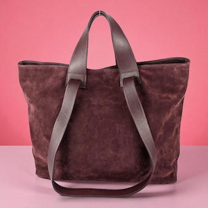 Op de afbeelding: Een bordeauxrode suède tote bag met bijpassende leren handvatten. De tas heeft een rechthoekige vorm en een ruim interieur. De handvatten zijn lang genoeg om over de schouder te dragen. De tas staat tegen een roze achtergrond.