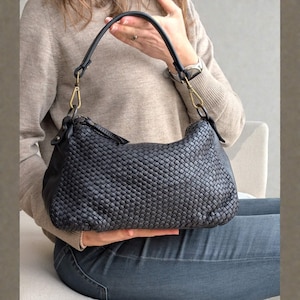 May include: A dark gray woven leather handbag with a curved shape. The bag has a black leather strap with gold-colored metal clasps. The bag is held by a person wearing a beige sweater and blue jeans.