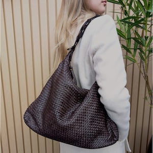 Könnte beinhalten: Eine dunkelbraune Hobo-Tasche mit einem komplexen geflochtenen Ledermuster auf der gesamten Außenfläche. Die Tasche verfügt über einen einzelnen Schulterriemen mit geflochtenem Design und eine lockere, unstrukturierte Form.