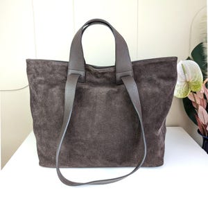 May include: A brown suede tote bag with matching brown leather handles. The bag has a rectangular shape and a soft, textured appearance. The handles are attached at the top and extend down the sides.