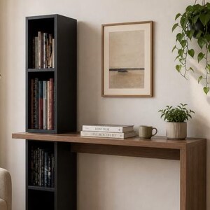 May include: A modern interior scene featuring a black bookshelf with books, a wooden desk, and a framed artwork. A small potted plant and a mug sit on the desk. A hanging plant and a lamp are also visible.