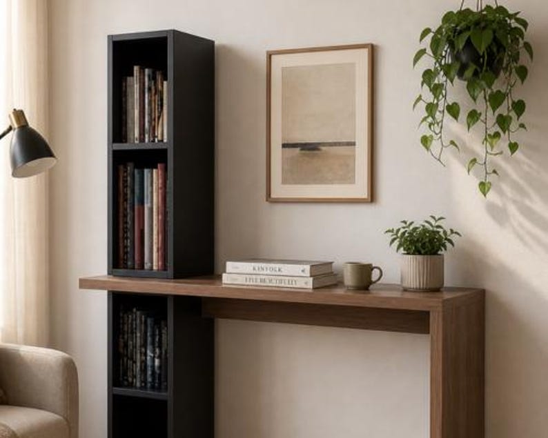 May include: A modern interior scene featuring a black bookshelf with books, a wooden desk, and a framed artwork. A small potted plant and a mug sit on the desk. A hanging plant and a lamp are also visible.