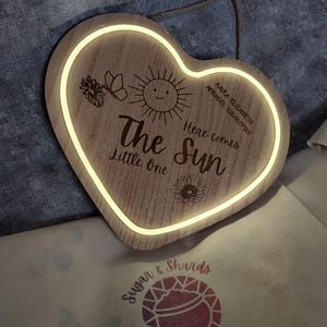 May include: Heart-shaped wooden sign with a glowing border. The sign features the text "The Sun Little One" with a smiling sun and flower illustrations. The sign also has the text "Here comes" and a name.