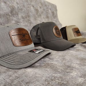 May include: Three trucker hats with leather patches. The first hat is gray with a black mesh back and a brown leather patch with the word "Skylane" and an airplane graphic. The second hat is gray with a brown leather patch. The third hat is tan and brown.