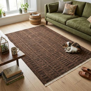 Puede incluir: Una alfombra rectangular marrón con un patrón geométrico de líneas horizontales. La alfombra está colocada sobre un suelo de madera en un salón. Un gato durmiente descansa sobre la alfombra cerca de un sofá verde. La alfombra tiene un borde con flecos.