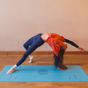 May include: A person in a yoga pose on a light blue yoga mat. The person is wearing navy blue leggings, an orange top, and white socks. The yoga mat has a circular design in the centre and lines along the edges.