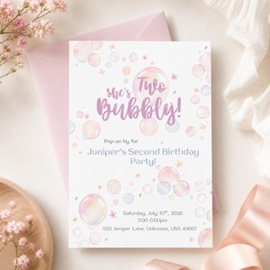 May include: A light pink invitation card with a matching envelope. The card reads "she's Two Bubbly!" and announces Juniper's Second Birthday Party. The design includes bubble illustrations and floral accents. The date and time are also included.