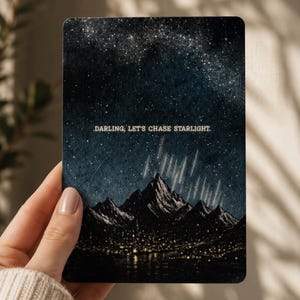 May include: A rectangular card featuring a dark night sky filled with stars and a meteor shower above a mountain range. The base of the mountains shows a glowing town near water. The text DARLING, LET'S CHASE STARLIGHT is printed in white.