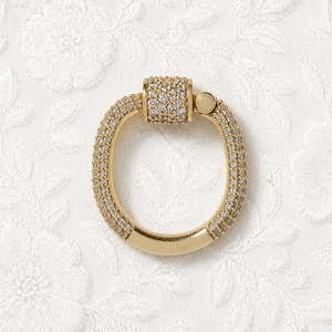 May include: A gold-colored, oval-shaped clasp with a hinged closure. The clasp is encrusted with small, clear gemstones. The background is a white fabric with a floral pattern.