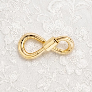 May include: A gold-colored infinity clasp, a jewelry component, lies on a white, floral-patterned fabric. The clasp is a polished metal with a smooth, reflective surface. The design is a continuous loop, with a central rectangular bar.