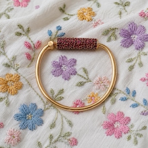 May include: A gold-toned bangle bracelet featuring a smooth, polished metal band and a cylindrical centerpiece encrusted with small, deep red crystals. The bracelet is circular in shape and has a hinged mechanism on one side of the crystal bar.
