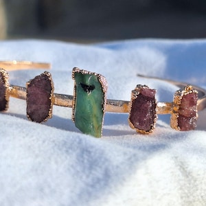 Op de afbeelding: Een goudkleurige manchetarmband met ruwe edelstenen. De armband heeft een centrale groene steen, geflankeerd door verschillende roze-paarse stenen. De edelstenen zijn gezet in goudkleurig metaal, wat een natuurlijk en elegant ontwerp creëert.