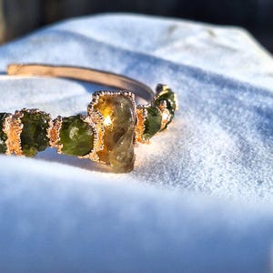 May include: A gold-toned cuff bracelet featuring a row of green and yellow-green gemstones. The bracelet has an open design and is resting on a white surface. The gemstones are irregularly shaped and set in gold-colored metal.