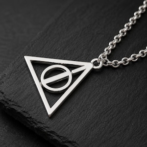 May include: A silver-toned metal pendant necklace featuring a geometric design of a triangle enclosing a circle with a vertical line bisecting it. The pendant is attached to a matching silver-toned metal chain with rounded, interlocking links.