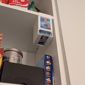 May include: A white cupboard interior with shelves containing food items. A box of tea bags labelled "Schwedische Blaubeere" is mounted on the shelf. Other items include a milk carton, a can of coffee, and a box of fruit-flavored tea.