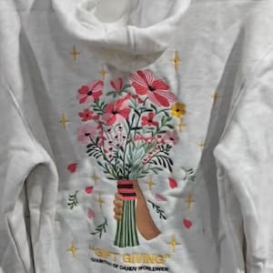 May include: A light gray hoodie with a floral embroidery design. The embroidery features a bouquet of pink, red, and yellow flowers held by a brown hand. The text "GIFT GIVING" is embroidered at the bottom.