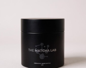 May include: A black cylindrical container with a matte finish. The text "THE MATCHA LAB" is printed in silver, along with the words "CEREMONIAL GRADE MATCHA" and "40G". The container is designed to hold matcha powder.