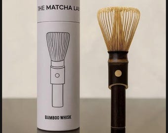May include: A bamboo matcha whisk with a dark handle and a bundle of thin tines. Beside it is a white cylindrical container with the text "THE MATCHA LAB" and a line drawing of a whisk. The text "BAMBOO WHISK" is below the drawing.