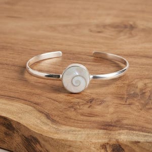 May include: A silver cuff bracelet featuring a circular centre piece inlaid with a white shiva shell. The shell displays a natural spiral pattern in shades of cream and grey. The metal band is smooth, polished, and open at the back for adjustment.