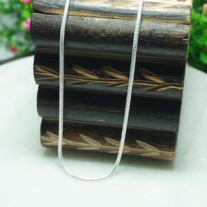 May include: A thin, silver-coloured chain necklace with a smooth, woven texture. The metallic chain features a consistent, interlocking link pattern that creates a sleek, reflective surface. The necklace is displayed in a simple, vertical loop.