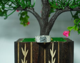 May include: A silver-colored ring with a textured, hammered finish. The band has a consistent width and a reflective, metallic surface that catches the light, showcasing a series of small, irregular indentations across the entire outer circumference.