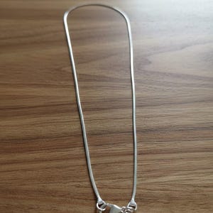 May include: A silver snake chain necklace with a lobster clasp. The chain is thin and flexible, and the clasp is a standard design. The necklace is displayed on a wooden surface.