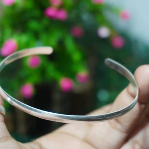 Könnte beinhalten: Ein schlichter Armreif aus silberfarbenem Metall mit glatter, polierter Oberfläche und offenem Design. Das Armband besteht aus einem dünnen, abgerundeten Band, das sich zu einer Kreisform biegt und eine saubere, reflektierende Oberfläche zeigt.