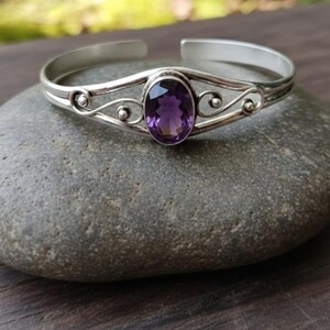 May include: Silver cuff bracelet with a deep purple oval amethyst gemstone. The bracelet has a decorative design with small, round accents. The bracelet is displayed on a gray stone. The amethyst is a beautiful, deep purple colour.