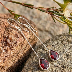 Op de afbeelding: Zilveren oorhangers met druppelvormige, gefacetteerde, dieprode edelstenen. De oorbellen worden getoond op een getextureerd, lichtbruin rotsachtig oppervlak, met groene en witte bloemaccenten op de achtergrond.