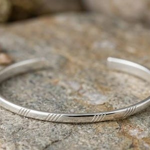 May include: A silver cuff bracelet with a simple, elegant design. The bracelet features three sets of parallel lines etched into the metal. The bracelet is displayed on a gray stone surface with small pebbles scattered around it.
