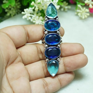 May include: A long silver ring featuring five vertically aligned faceted gemstones in shades of blue. The top and bottom stones are pear-shaped light blue, while the three center stones are oval-shaped in varying shades of deep royal blue.