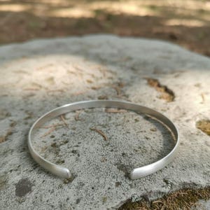 Może przedstawiać: Srebrna, otwarta bransoletka typu cuff. Bransoletka wykonana jest z cienkiej, zaokrąglonej metalowej opaski o gładkim, matowym wykończeniu. Końce bransoletki są lekko zaokrąglone. Bransoletka spoczywa na szarej kamiennej powierzchni.