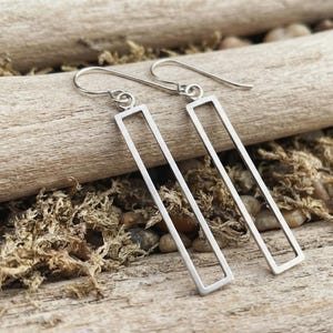 May include: A pair of silver-toned earrings featuring an open rectangular frame design. Each earring hangs from a curved hook wire, showcasing a clean, geometric silhouette with a smooth metallic finish and a hollow center structure.