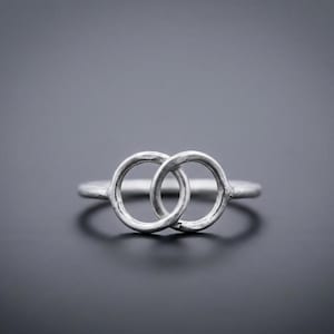 Può includere: Un anello d'argento con due cerchi intrecciati. L'anello è in metallo chiaro e poggia su una superficie grigio scuro. I cerchi hanno una texture e l'anello è a fuoco.