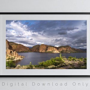 Könnte beinhalten: Gerahmte Landschaftsfotografie mit einem See, umgeben von Bergen unter einem dramatischen, bewölkten Himmel. Das Bild zeigt grüne Vegetation und Kakteen im Vordergrund. Der Text unten lautet "Digital Download Only".