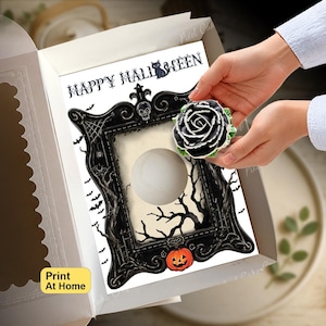 May include: A white cardboard box containing a rectangular card with the text HAPPY HALLOWEEN, a black ornate frame illustration with a skull, a pumpkin, and a circular cutout, and a black rose cupcake topper with green leaves held by hands.