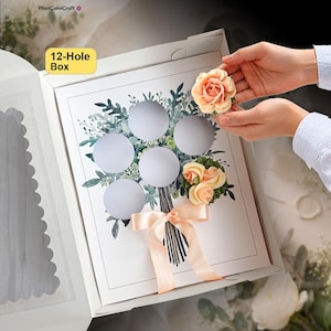 May include: A white 12-hole cupcake box featuring a printed bouquet illustration with green foliage and a peach-coloured ribbon. The box includes circular cutouts for cupcakes, with two peach-coloured frosting flowers placed on the insert.