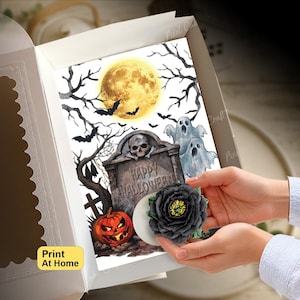 May include: A rectangular card featuring a Halloween illustration with a full moon, bare trees, bats, two ghosts, a tombstone with the text Happy Halloween, and a jack-o'-lantern, partially covered by a dark gray artificial flower.
