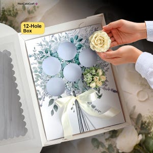 May include: A white 12-hole cupcake box featuring a printed bouquet of green leaves and stems. The box includes a cream-colored ribbon tied in a bow at the bottom. A single cream-colored rose-shaped cupcake is being placed into one of the holes.