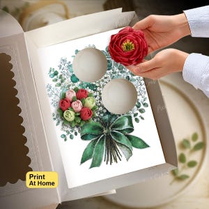 May include: A white cupcake box insert featuring a printed illustration of a floral bouquet with red and green frosting-style flowers, a large green bow, and two circular cutouts for cupcakes, with a red flower held above the insert.