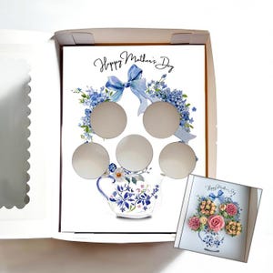 May include: A white box with a floral design and the text "Happy Mother's Day". The design includes a blue bow, flowers, and a teapot. The box has five circular cutouts. A smaller card with a floral arrangement is also visible.