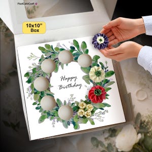 Può includere: Una scatola bianca di 25,4x25,4 cm contenente un cartoncino quadrato con un'illustrazione ad acquerello di una corona di foglie verdi. Il cartoncino riporta la scritta Happy Birthday in corsivo nero e include cinque sfere di legno e tre fiori in tessuto.