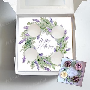 May include: A white box containing a square cake decorated with a lavender wreath and the words "Happy Birthday." The cake has four circular cutouts. A smaller card with a similar design and floral accents is also visible.