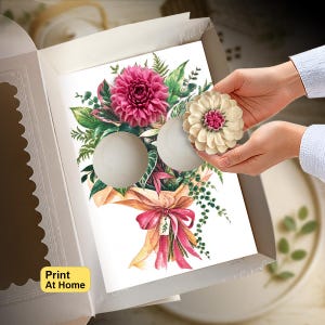 May include: A white cardboard box containing a printed sheet with a floral bouquet illustration featuring a large pink dahlia, green foliage, and a tan paper wrap with a pink ribbon. A cream-colored flower-shaped cupcake topper is being placed.