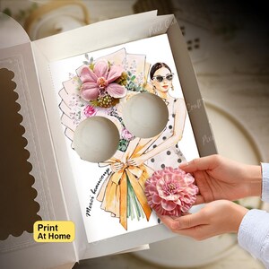 May include: A white cardboard box containing a printed card featuring an illustration of a person holding a large bouquet of flowers, with two circular cutouts for cupcakes and the text Merci beaucoup, accompanied by a pink decorative flower.