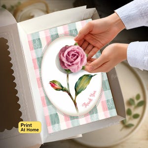 Könnte beinhalten: Eine rechteckige Karte mit einer rosa Rosenillustration in einem ovalen Rahmen auf einem rosa-grün karierten Hintergrund. Die Karte enthält den Text Thank You und ein gelbes Feld mit dem Text Print At Home. Die Karte liegt in einer weißen Schachtel.