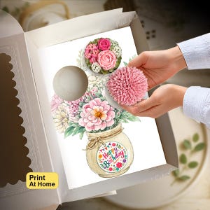 May include: A white cardboard box containing a printed card with a watercolor illustration of a beige vase filled with pink and white flowers. A round tag on the vase reads Happy Birthday. A pink textured pom-pom is held over the card illustration.