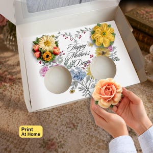 Puede incluir: Un inserto de cartón blanco para caja de cupcakes con un diseño floral y el texto Happy Mother's Day. El inserto tiene dos aberturas circulares y está decorado con flores estilo acuarela en amarillo, rosa y azul. Se sostiene una rosa de glaseado color melocotón.