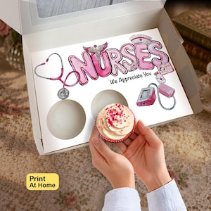 以下が含まれることがあります： NURSESとWe Appreciate Youというピンクの文字が書かれた白いカップケーキ用ボックスの仕切り。聴診器、ナースキャップ、医療用バッジ、薬瓶、血圧計のイラストが描かれており、カップケーキが1つ入っています。